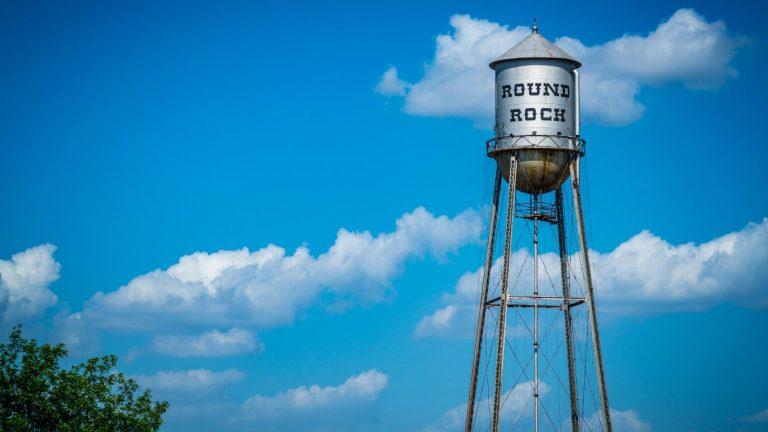 An image of a large, metal water tower with “ROUND ROCK” painted on it, standing against a clear blue sky. Best Places to Live in Round Rock Texas.